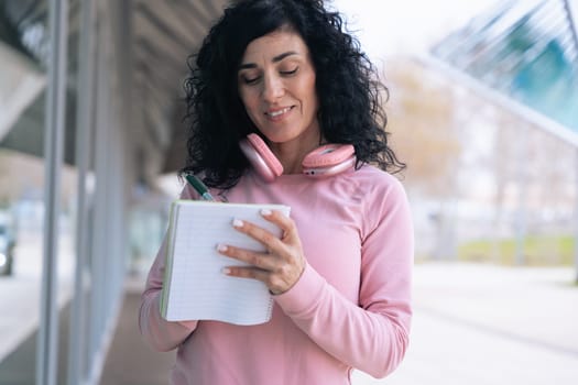 Caucasian sporty woman in a pink hoodie writing in a notebook outdoors. Wearing headphones around her neck, she appears focused and relaxed after a workout in an urban setting.