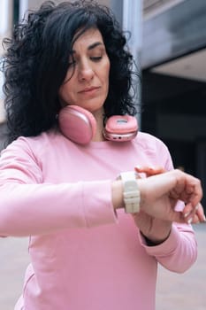 Middle adult Caucasian woman with curly black hair checking her smartwatch outdoors. She is wearing a pink sweatshirt and pink headphones, representing fitness, technology, and an active lifestyle.