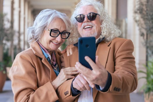 Happy caucasian senior couple taking a selfie with a smartphone. They smile brightly, wearing sunglasses and jackets. Concept of love, joy, connection, and modern lifestyle in an outdoor setting.