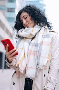 Serious caucasian woman standing outdoors in urban setting, wearing coat and scarf, using her red smartphone. She is serious and relaxed, enjoying a casual moment on a cold day in the city. Vertical