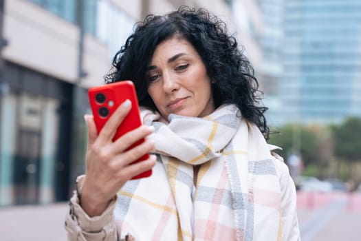 Serious caucasian woman standing outdoors in urban setting, wearing coat and scarf, using her red smartphone. She is serious and relaxed, enjoying a casual moment on a cold day in the city.