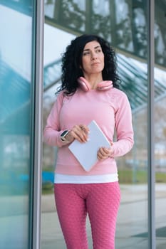 Caucasian sporty woman in pink activewear holding a notebook and pen. Standing outside near a glass building with headphones around her neck, she looks thoughtful and focused after training.