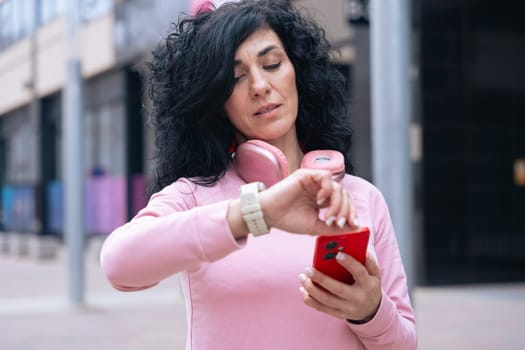Middle adult Caucasian woman with curly black hair checking her smartwatch outdoors. She is wearing a pink sweatshirt and pink headphones, representing fitness, technology, and an active lifestyle.