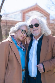 Caucasian elderly couple standing close in a sunny park. The woman leans her head on the man shoulder, both wearing brown jackets and glasses, sharing a quiet and affectionate moment.Vertical