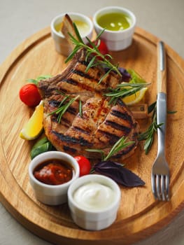 A beautifully presented grilled pork chop sits on a wooden platter, accompanied by fresh herbs, colorful vegetables, and a variety of sauces. Perfect for a hearty meal.