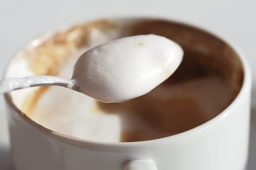 A spoon with rich foam hovers above a warm coffee cup on a table.
