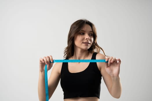 Brunette woman in black t-shirt holding a centimeter tape in front of her face with a grey background. Sport, fitness and diet