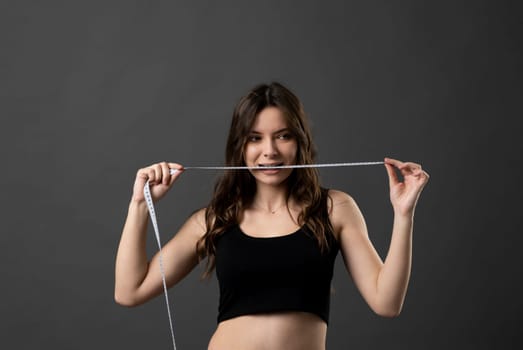 Angry brunette woman in black t-shirt holding a centimeter tape in a mouth with a grey background. Sport, fitness and diet