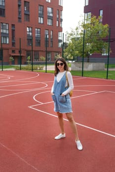 A young woman in a denim dress stands on a red outdoor basketball court, smiling confidently.