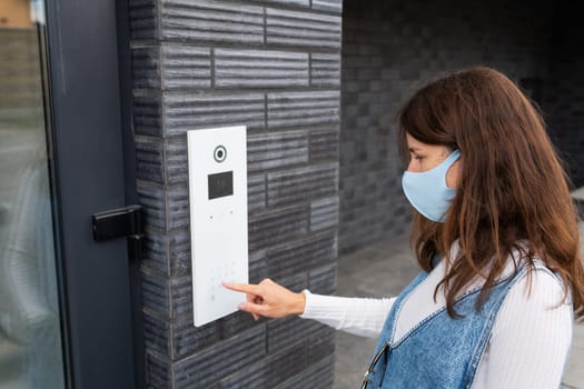 A young woman wearing a face mask uses a digital door intercom outside a modern building.