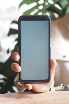 A person displays a blank smartphone screen against lush plants indoors.