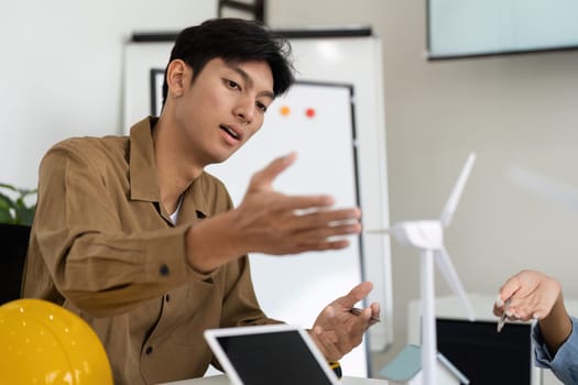 In a collaborative workspace, a young professional explains the benefits of wind energy while pointing at a miniature wind turbine model, signaling innovation in sustainable design.