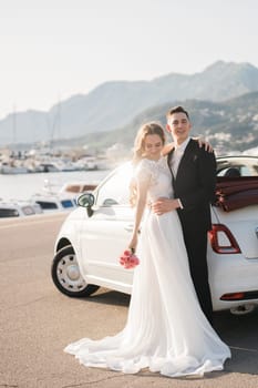 Stylish newlyweds are hugging near a white car in a green park, against the backdrop of summer nature. Bearded groom and blond bride in a lace dress are smiling outdoors. Wedding portrait.