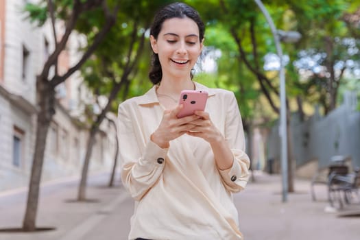 Young caucasian woman smiling while using her smartphone on a tree-lined urban street. She looks relaxed and engaged with her device during a pleasant outdoor walk in the city.