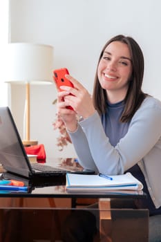 Smiling Caucasian woman holding smartphone and looking at camera. Sitting at home office with laptop, notebook, and decor. Modern workspace, technology, communication, education, and productivity.