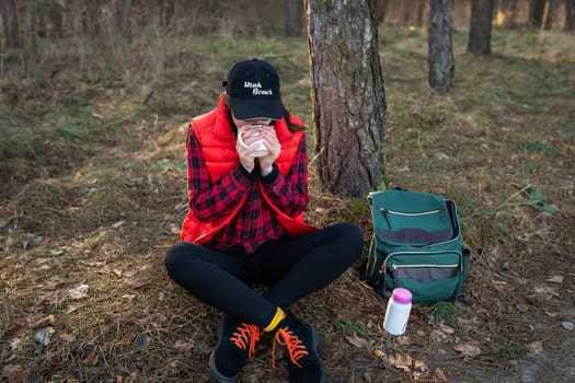 Trekking, hiking. A backpack with a thermos and a cup stands near a tree, a girl warms herself with hot tea, a snack in the open air.Travel and tourism