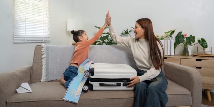 In a cozy living room, a mother and daughter enjoy a fun moment, high-fiving over their travel plans while sitting beside their suitcase.