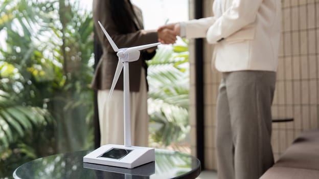 A close-up on two women shaking hands, with a miniature wind turbine in the foreground, symbolizing their commitment to sustainable business practices.