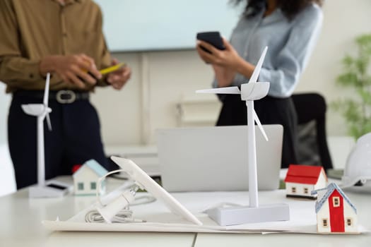 Two professionals engage in a collaborative meeting, brainstorming innovative sustainable energy solutions using miniature models of wind turbines and eco-friendly houses.