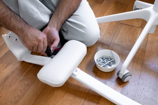 Male adult assembling white office chair with tools and screws on wooden floor.