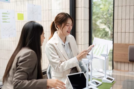 Two women engage in a productive discussion about sustainable business strategies, reviewing plans and ideas in a modern office filled with greenery.