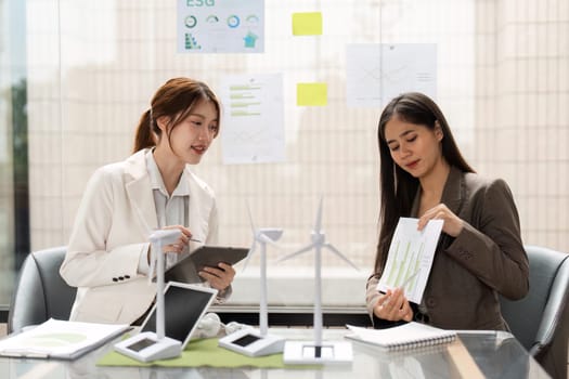 In a sleek office, two women discuss ESG strategies while reviewing charts and models, emphasizing sustainable solutions and data-driven decisions for better environmental impact.