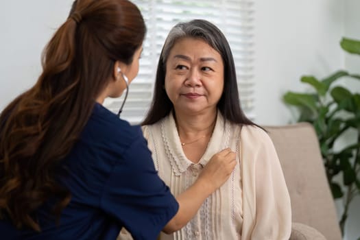 A caregiver uses a stethoscope to listen to the heart of an elderly patient sitting comfortably at home, emphasizing the importance of regular health check-ups and personal connection.