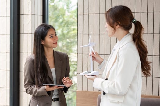 Two women engage in a lively conversation about sustainability and renewable energy, showcasing innovative solutions through a tablet and a model wind turbine.