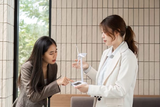 In a sleek workplace, two women engage in a conversation about sustainable technology, showcasing a miniature wind turbine model as they strategize advancements in ESG initiatives.
