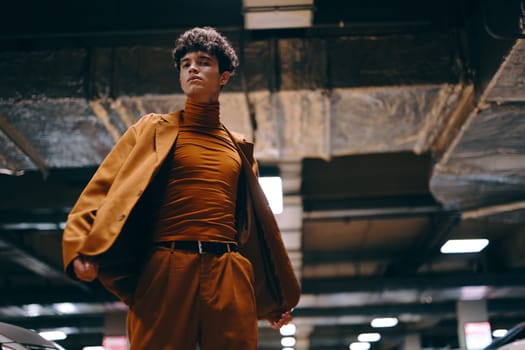 A fashionable young man stands confidently, showcasing a brown turtleneck and blazer in a modern, industrial space