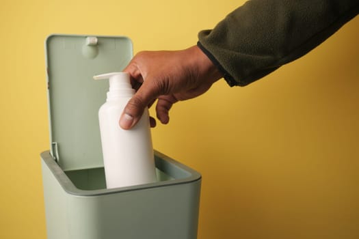 A hand drops a white bottle into a green trash can on yellow.