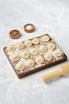Dough dumplings with beef on a wooden board, dusted with flour. A rolling pin and bowls of spices surround the dumplings, hinting at a homemade cooking experience in a kitchen setting.