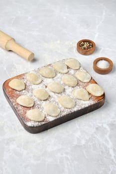 Homemade dumplings are neatly arranged on a floured wooden board. Nearby, a rolling pin and small bowls of salt and pepper add to the cooking preparation atmosphere.
