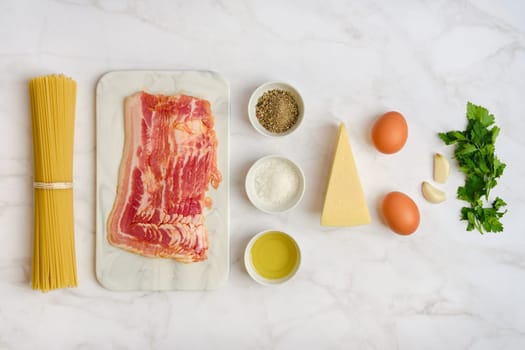 Fresh ingredients on a marble countertop for cooking spaghetti carbonara. Includes bacon, eggs, cheese, garlic, herbs, black pepper, and olive oil, ready for a delicious meal.