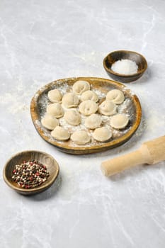 Dumplings on a wooden plate, sprinkled with flour, along with small bowls of salt and mixed pepper. A rolling pin rests beside them, highlighting the preparation process.