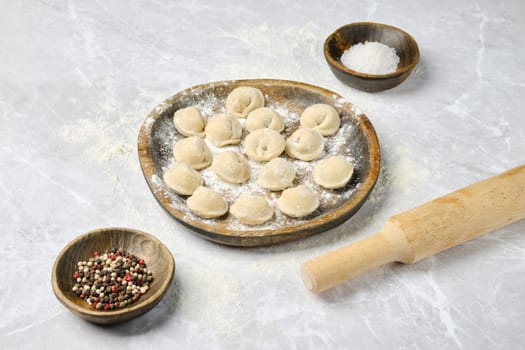 A wooden plate holds freshly made dumplings surrounded by bowls of spices and salt on a flour-dusted countertop. A rolling pin rests nearby, indicating the process of making these treats.