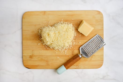 Shredded cheese on a wooden cutting board alongside a block of cheese and a grater. The items are arranged neatly in a well-lit kitchen, suggesting preparation for a delicious meal.