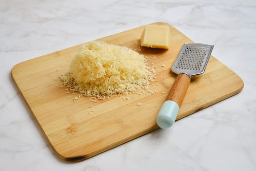 A wooden cutting board holds a pile of finely shredded cheese beside a block of cheese and a metal grater. The items are arranged neatly, showcasing a kitchen preparation setup.