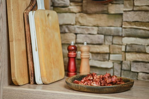 A rustic kitchen features a plate of chopped bacon next to wooden cutting boards. Two shakers, one wooden and one red, sit in the background, adding to the warm atmosphere.