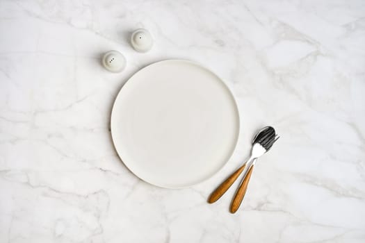 A simple dining arrangement showcases an empty white plate accompanied by a fork and knife with wooden handles, placed on a smooth marble surface, alongside salt and pepper shakers.