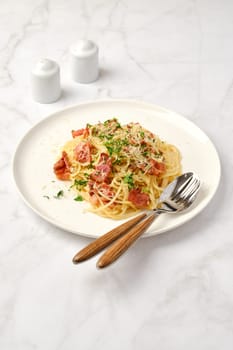 Spaghetti carbonara topped with crispy bacon pieces and grated cheese sits on a white plate. Two forks are placed beside the dish, and condiments are seen in the background.