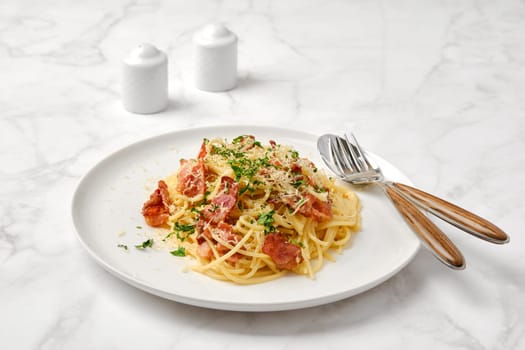 A delicious plate of spaghetti carbonara topped with crispy bacon, grated cheese, and fresh herbs on a marble table. Small salt and pepper shakers complete the casual dining setting.