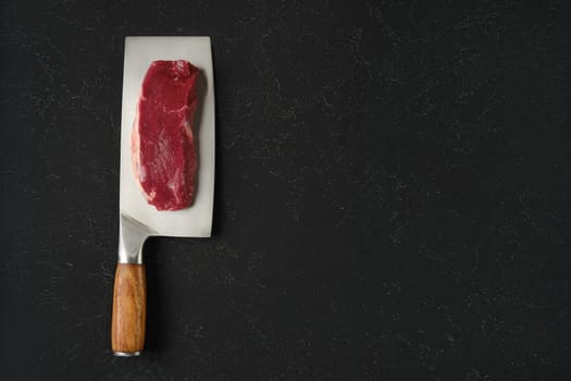 A piece of raw beef meat on a sharp cleaver with a wooden handle. The cleaver rests on a black countertop, showcasing the texture of the meat and the gleam of the blade.