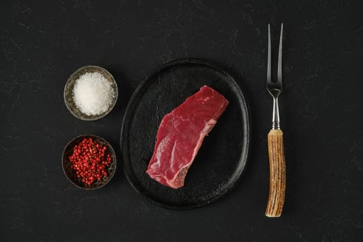 A carefully portioned piece of raw beef is placed on a black plate surrounded by bowls of pink peppercorn and salt, with a rustic fork showcasing a ready-to-cook atmosphere.