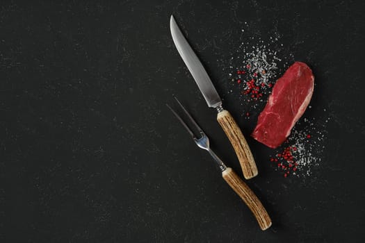 A knife and fork, with horn handles, rest beside a raw beef steak on a dark surface. Salt and pink peppercorn surround the meat, suggesting preparation for cooking or a gourmet meal.