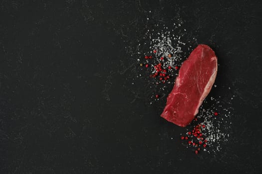 A piece of fresh raw beef sirloin steak on a dark countertop surrounded by coarse sea salt and red peppercorns, showcasing a clean and elegant presentation ideal for meal preparation.