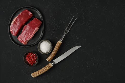 Two cuts of raw beef steak on a plate alongside small bowls with salt, pink peppercorns, and tools for cutting. The setting highlights a refined culinary preparation.