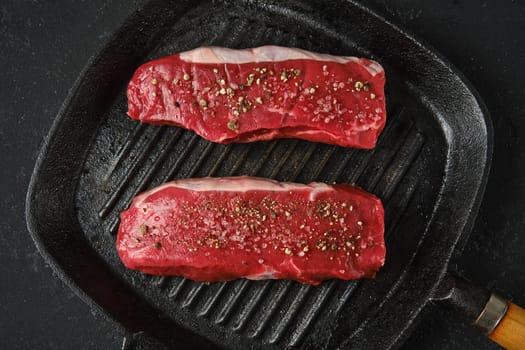 Top view of two thick cuts of raw beef steaks on a cast iron skillet. The marbled texture of the meat is seasoned with sea salt and pepper