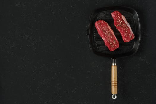 Two raw beef steaks on a preheated griddle pan, seasoned with spices, in a home kitchen setting. The black surface is for text