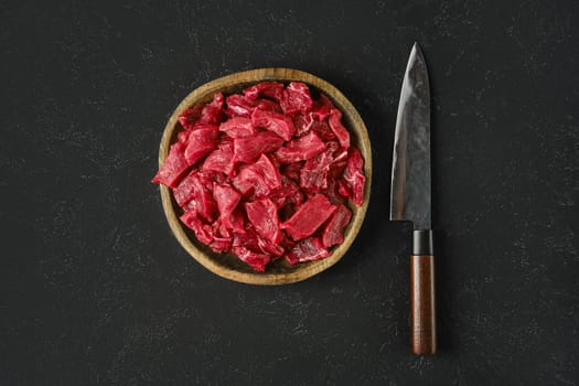 A wooden bowl holds freshly cut pieces of raw beef, showcasing a vibrant red color. Beside the bowl, a sharp knife rests on a dark textured surface, ready for preparation or cooking.